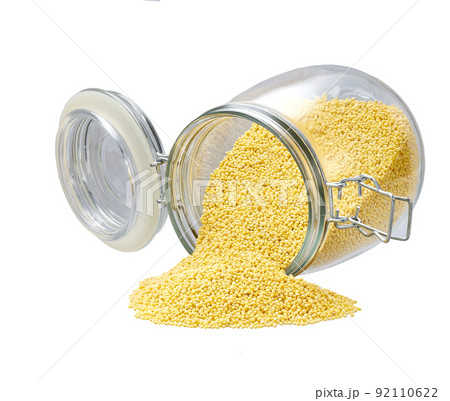 Raw grains of millet spill out of a glass storage jar isolated on white background. 92110622