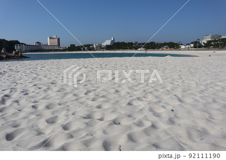 白い砂浜の砂にピントを合わせ海と海岸と木々の森と宿泊施設の建物と青空の見える風景 92111190