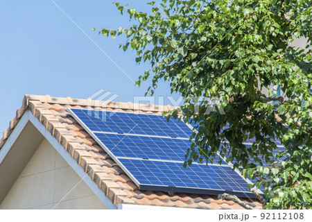 【環境イメージ】太陽光パネルが設置された住宅の屋根と快晴の青空と新緑の葉っぱ。 92112108