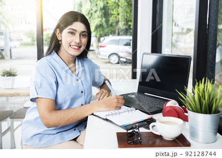 Asian woman writing something on a notebook with a laptop and a cup of coffee on the table 92128744