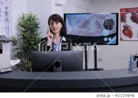 Portrait of smiling hospital receptionist working at front desk using telephone talking with patient making an appointment. Professional looking woman answering phone call at private clinic. 92133648
