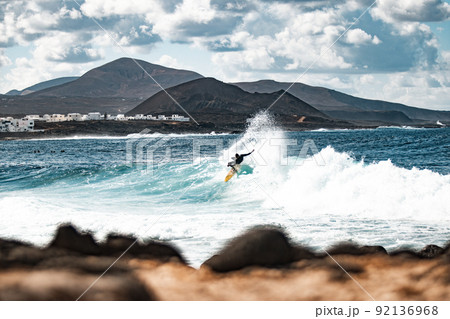 Wild rocky coastline of surf spot La Santa Lanzarote, Canary Islands, Spain. Surfer riding a big wave in rocky bay, volcano mountain in background. Wild rocky coastline of surf spot La Santa Lanzarote, Canary Islands, Spain. Surfer riding a big wave in rocky bay, volcano mountain in background. 92136968