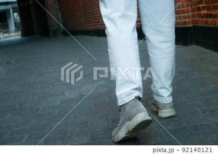 A woman in jeans walks through the narrow streets of the old city. Cropped photo back view of legs. Brick walls around the pedestrian alley. 92140121