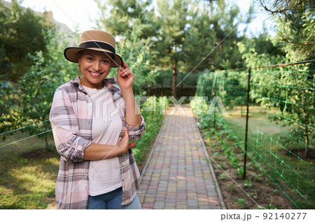 Pleasant pretty Hispanic woman, female gardener smiles looking at camera, against an organic vegetable garden background Pleasant pretty Hispanic woman, female gardener smiles looking at camera, against an organic vegetable garden background 92140277