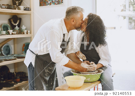 Mutual creative work. Adult elegant couple in casual clothes and aprons. People creating a bowl on a pottery wheel in a clay studio. Mutual creative work. Adult elegant couple in casual clothes and aprons. People creating a bowl on a pottery wheel in a clay studio. 92140553