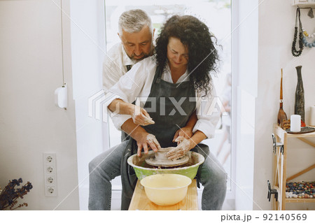 Mutual creative work. Adult elegant couple in casual clothes and aprons. People creating a bowl on a pottery wheel in a clay studio. Mutual creative work. Adult elegant couple in casual clothes and aprons. People creating a bowl on a pottery wheel in a clay studio. 92140569