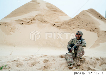Military or soldier man with sunglasses sit on sand and look around in area look like desert Military or soldier man with sunglasses sit on sand and look around in area look like desert 92141284