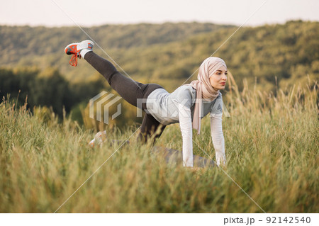 Smiling young woman in hijab and sport outfit doing flexible exercises on yoga mat Smiling young woman in hijab and sport outfit doing flexible exercises on yoga mat 92142540