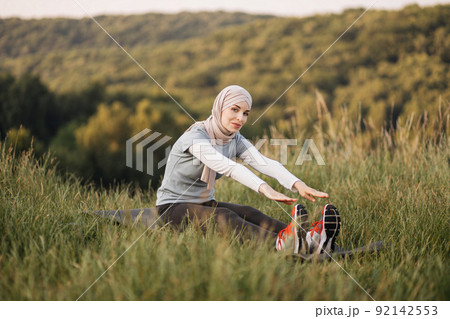 Pleasant woman in hijab and sport clothes sitting on yoga mat and stretching body. Pleasant woman in hijab and sport clothes sitting on yoga mat and stretching body. 92142553