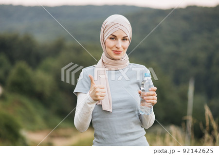 Muslim woman in hijab and sport clothes standing at green park and holding bottle of water. 92142625