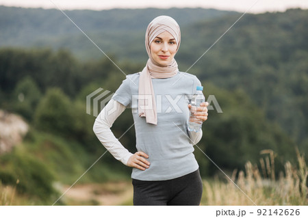 Muslim woman in hijab and sport clothes standing at green park and holding bottle of water. Muslim woman in hijab and sport clothes standing at green park and holding bottle of water. 92142626