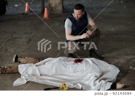 Young man in bulletproof vest sitting on squats by covered dead body 92143094