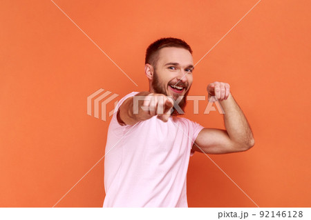Portrait of handsome bearded man pointing to camera, smiling, making happy choice, we need you concept, wearing pink T-shirt. Indoor studio shot isolated on orange background. 92146128