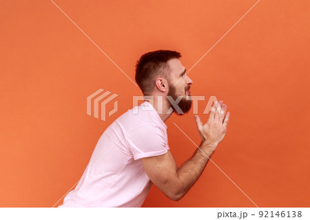 Side view of bearded man folding hands in prayer, closing eyes and talking to god, asking for help, expressing gratitude, wearing pink T-shirt. Indoor studio shot isolated on orange background. Side view of bearded man folding hands in prayer, closing eyes and talking to god, asking for help, expressing gratitude, wearing pink T-shirt. Indoor studio shot isolated on orange background. 92146138