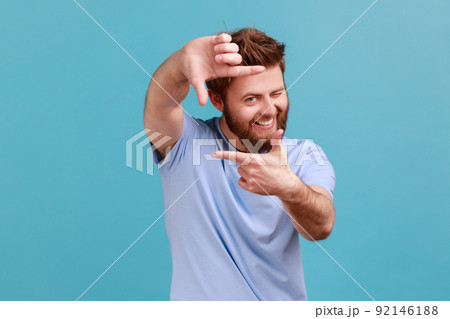 Portrait of curious bearded man gesturing picture frame with hands, looking through fingers and focusing on interesting moment, taking photo. Indoor studio shot isolated on blue background. 92146188