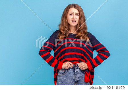 Upset woman wearing striped casual style sweater, trying to close the buttons of her jeans, gained extra weight during the holidays. Indoor studio shot isolated on blue background. Upset woman wearing striped casual style sweater, trying to close the buttons of her jeans, gained extra weight during the holidays. Indoor studio shot isolated on blue background. 92146199