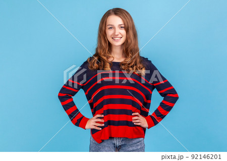 Portrait of smiling positive woman wearing striped casual style sweater looking at camera, keeping hands on hips, having confident expression. Indoor studio shot isolated on blue background. 92146201