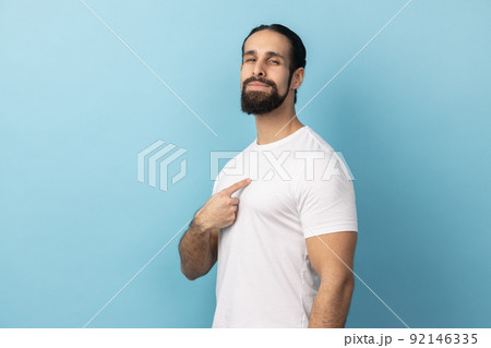 Portrait of self confident man with beard wearing white T-shirt pointing at himself, feeling proud and self-important, having big ego. Indoor studio shot isolated on blue background. 92146335