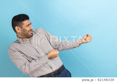 Side view of strong purposeful businessman with beard pulling invisible rope, showing his persistence and leadership qualities, wearing striped shirt. Indoor studio shot isolated on blue background. Side view of strong purposeful businessman with beard pulling invisible rope, showing his persistence and leadership qualities, wearing striped shirt. Indoor studio shot isolated on blue background. 92146339