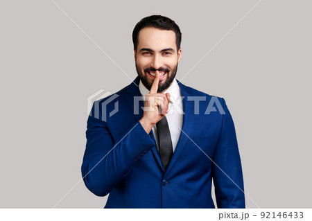 Smiling positive bearded man holding finger near lips showing shh gesture, keeping secrets preparing surprise, wearing official style suit. Indoor studio shot isolated on gray background. Smiling positive bearded man holding finger near lips showing shh gesture, keeping secrets preparing surprise, wearing official style suit. Indoor studio shot isolated on gray background. 92146433