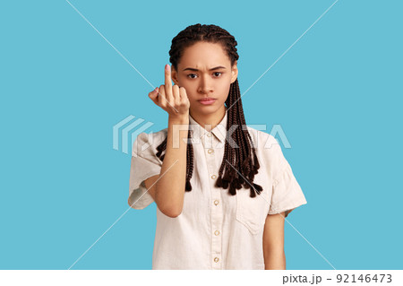 Serious woman with black dreadlocks demonstrates middle finger, looks with poker face, being vulgar and has quarrel with someone, wearing white shirt. Indoor studio shot isolated on blue background. 92146473
