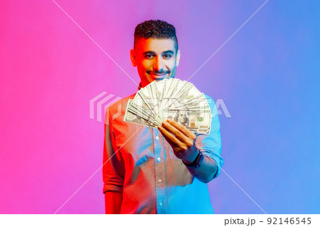 Portrait of rich handsome man in shirt holding dollars, looking at camera with toothy smile, being happy to win lottery. Indoor studio shot isolated on colorful neon light background. Portrait of rich handsome man in shirt holding dollars, looking at camera with toothy smile, being happy to win lottery. Indoor studio shot isolated on colorful neon light background. 92146545