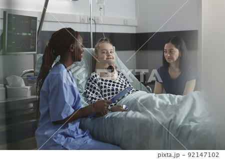 Unwell suffering kid recovering in pediatric hospital ward while resting in patient bed. Nurse examining injured girl with fractured neck wearing cervical collar because of car accident. Unwell suffering kid recovering in pediatric hospital ward while resting in patient bed. Nurse examining injured girl with fractured neck wearing cervical collar because of car accident. 92147102