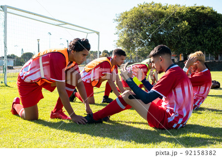 Male players holding legs of multiracial teammates practicing crunches on grassy field at playground 92158382