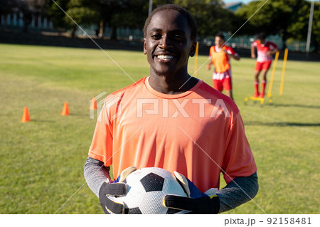 Portrait of african american smiling goalkeeper wearing gloves holding soccer ball at playground 92158481