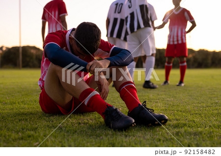 Caucasian sad male athlete sitting on grassy land with team players in background at playground 92158482