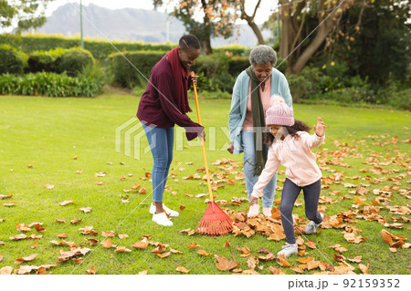 Image of happy african american grandmother, mother and daughter swiping leaves 92159352