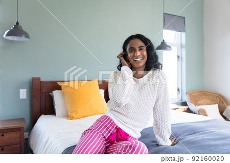 Portrait of cheerful biracial young woman smiling while relaxing on bed at home, copy space 92160020