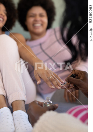 Hands of biracial young woman painting female friend's fingernails while spending leisure time 92160068