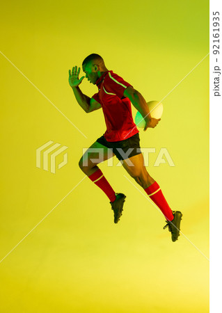 African american male rugby player running with rugby ball over yellow lighting 92161935