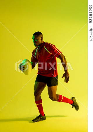 African american male rugby player running with rugby ball over yellow lighting African american male rugby player running with rugby ball over yellow lighting 92161939