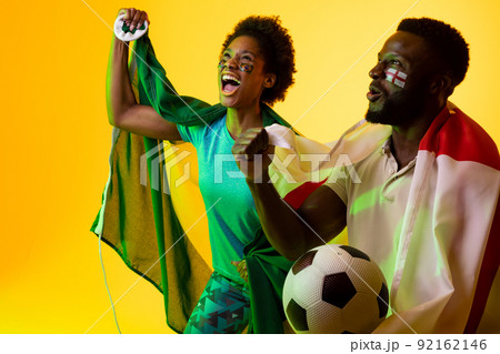 Image of african american soccer fan couple with flags of brazil and england in yellow lighting Image of african american soccer fan couple with flags of brazil and england in yellow lighting 92162146