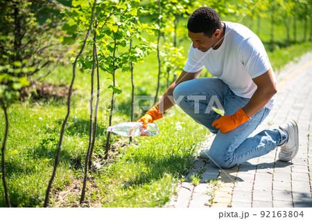 Man crouched watering small tree in park Man crouched watering small tree in park 92163804