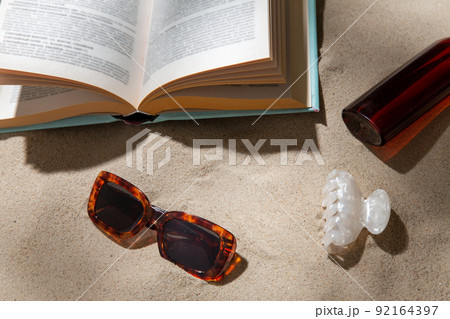 book, sunglasses and sunscreen on beach sand 92164397