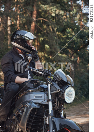 A young male biker travels on a motorcycle alone, stopped and puts on a helmet on the side of a forest road 92173158