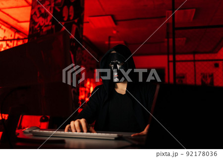 Dangerous unrecognizable hacker man wearing in black mask and sweatshirt hacking online web site using laptop and desktop, sitting at table in dark basement room with red neon lights. 92178036