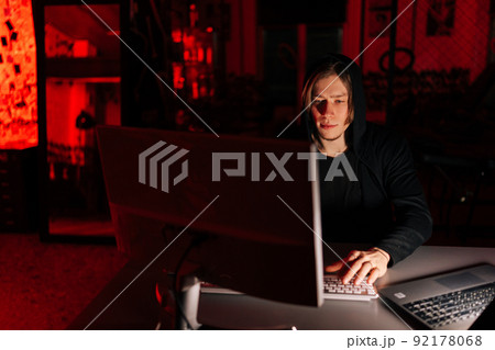 Portrait of wanted hacker man stealing information, preparing to break password sitting at desk in dark room with red neon lights. Concept of data thief, internet attack, darknet and cyber security. 92178068