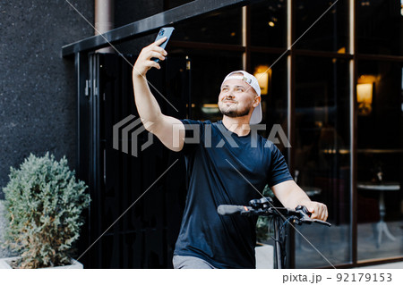 Adult man well dressed with electric scooter taking a selfie with his mobile phone in the street 92179153