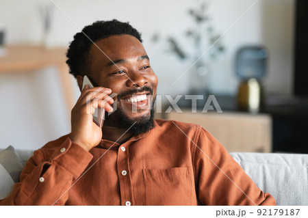 Cheerful Black Guy Talking On Cellphone Sitting On Sofa Indoor 92179187