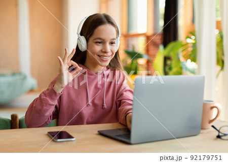 Studentgirl having online video class via laptop and gesturing OK, approving e-learning, sitting at desk and smiling Studentgirl having online video class via laptop and gesturing OK, approving e-learning, sitting at desk and smiling 92179751