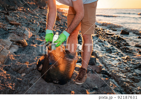 A male volunteer puts a plastic bottle in a bag full of garbage. In the background wild beach and ocean. Close up. The concept of environmental conservation 92187631