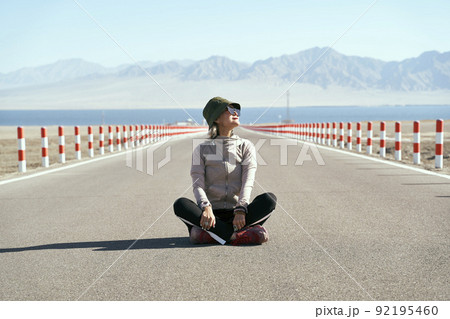 asian woman tourist sitting in the middle of an empty open road looking at view with lake and rolling mountains in background, leg crossed 92195460