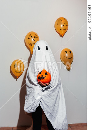 Happy Halloween. Ghost is holding orange pumpkin in hand. Child in white suit, painted yellow balloons. Different emotions of joy, anger, laughter. Festive design, party concept. Soft Selective focus Happy Halloween. Ghost is holding orange pumpkin in hand. Child in white suit, painted yellow balloons. Different emotions of joy, anger, laughter. Festive design, party concept. Soft Selective focus 92195463