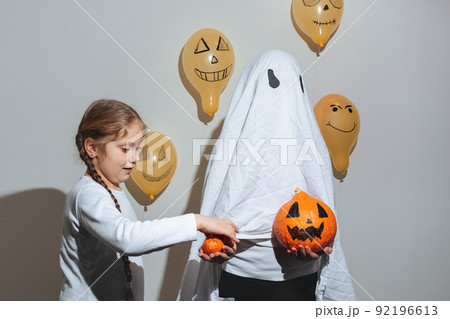 Happy Halloween. Girl gives ghost an orange pumpkin. Child in white suit. Painted yellow balloons. Different emotions of joy, anger, laughter. Festive design, party concept. Soft selective focus Happy Halloween. Girl gives ghost an orange pumpkin. Child in white suit. Painted yellow balloons. Different emotions of joy, anger, laughter. Festive design, party concept. Soft selective focus 92196613