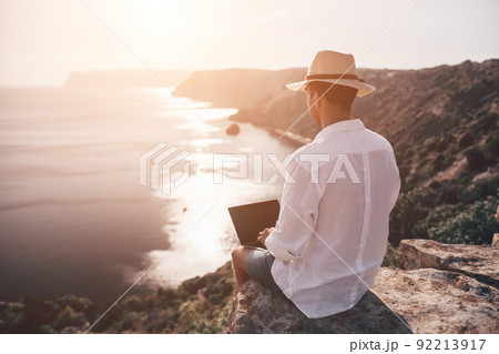 Digital nomad, freelancer. Remote work on vacation. Man in the hat, a businessman with a laptop sits on the rocks by the sea during sunset, makes a business transaction online from a distance. Digital nomad, freelancer. Remote work on vacation. Man in the hat, a businessman with a laptop sits on the rocks by the sea during sunset, makes a business transaction online from a distance. 92213917