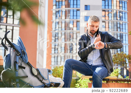 Father making business call, looking on watch while spending time with son outdoors. Front view of bearded man holding mobile phone, calling, while baby sleeping in pram in park. Concept of working. 92222689
